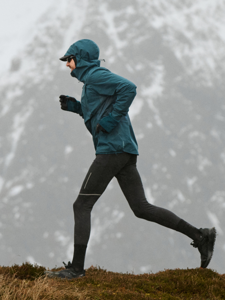 A person in a blue hooded jacket and black leggings runs along a grassy ridge with snowy mountains in the background.