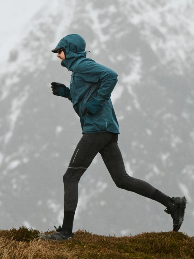 A person in a blue hooded jacket and black leggings runs along a grassy ridge with snowy mountains in the background.