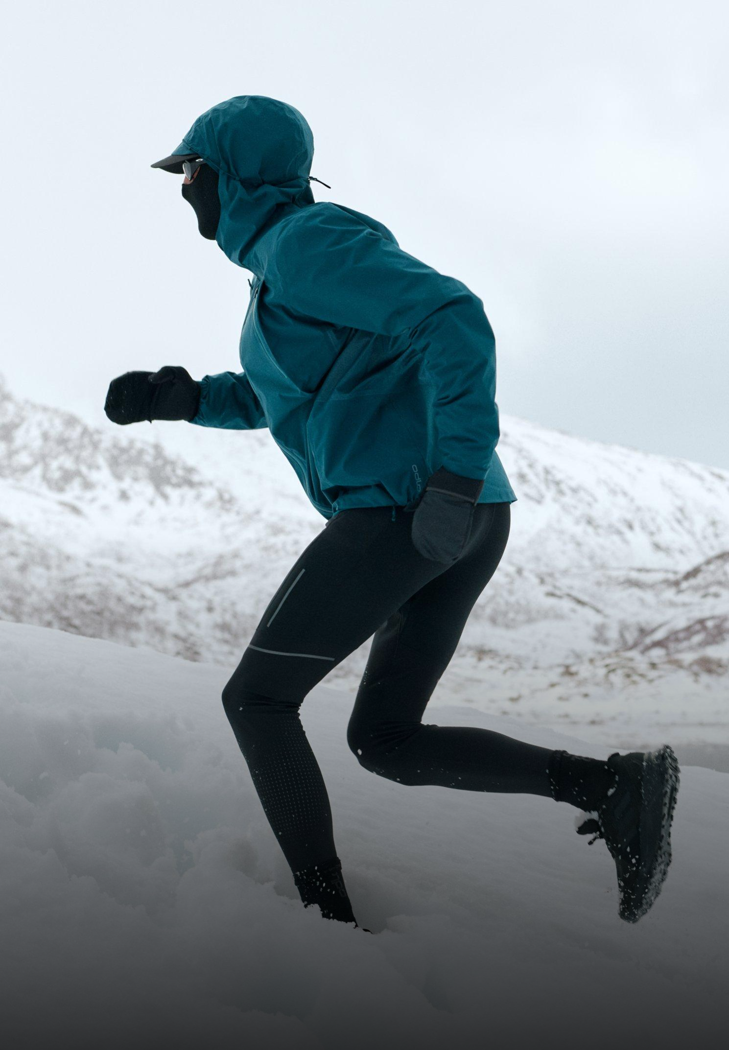 Persoon die bergop rent door diepe sneeuw, gekleed in een blauwgroene jas met capuchon en zwarte legging, in een besneeuwd berglandschap.