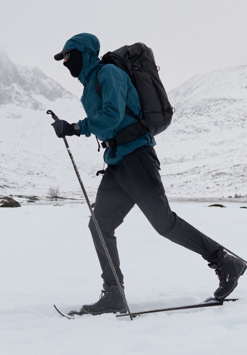Eine Person beim Skilanglauf in verschneiten Bergen, mit blauer Jacke, schwarzer Hose, Gesichtsmaske und schwarzem Rucksack.
