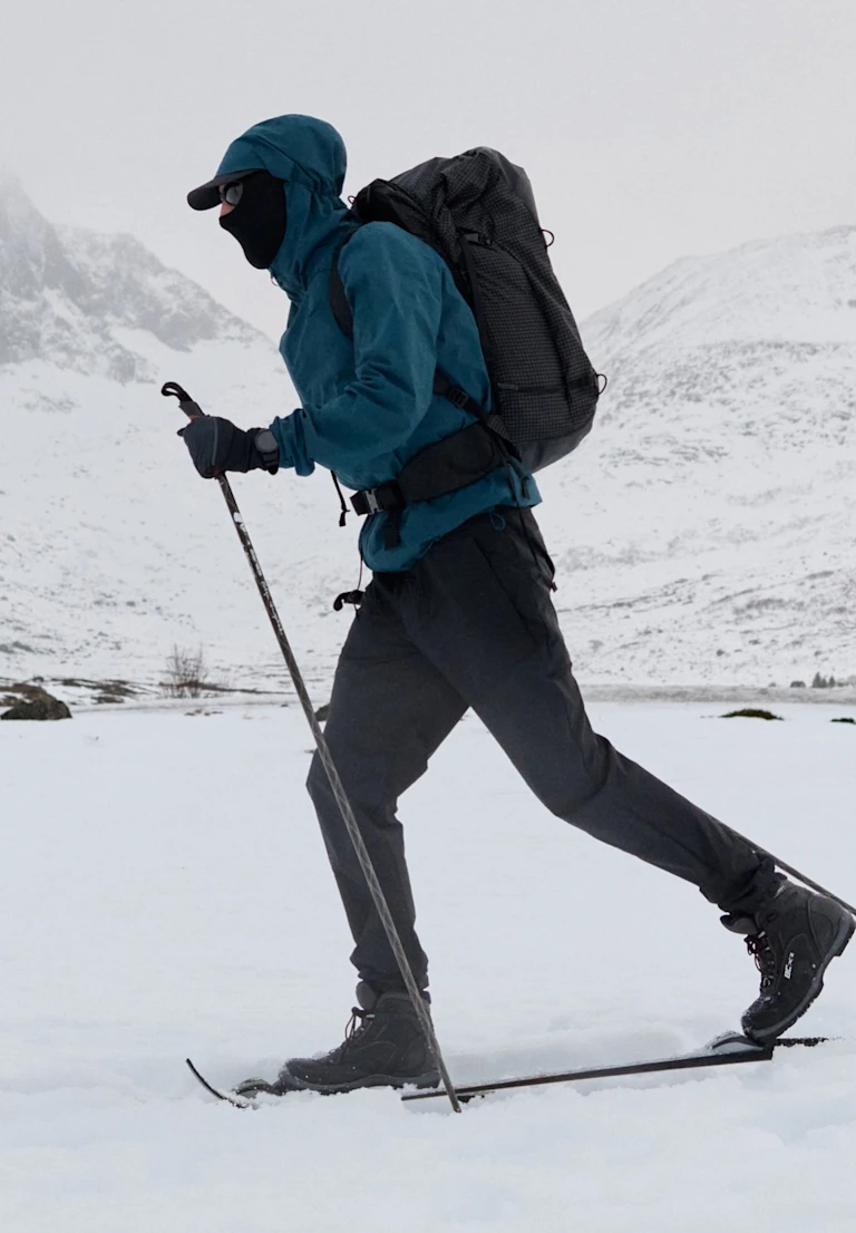 A person cross-country skiing in snowy mountains, wearing a blue jacket, black pants, face mask, and black backpack.