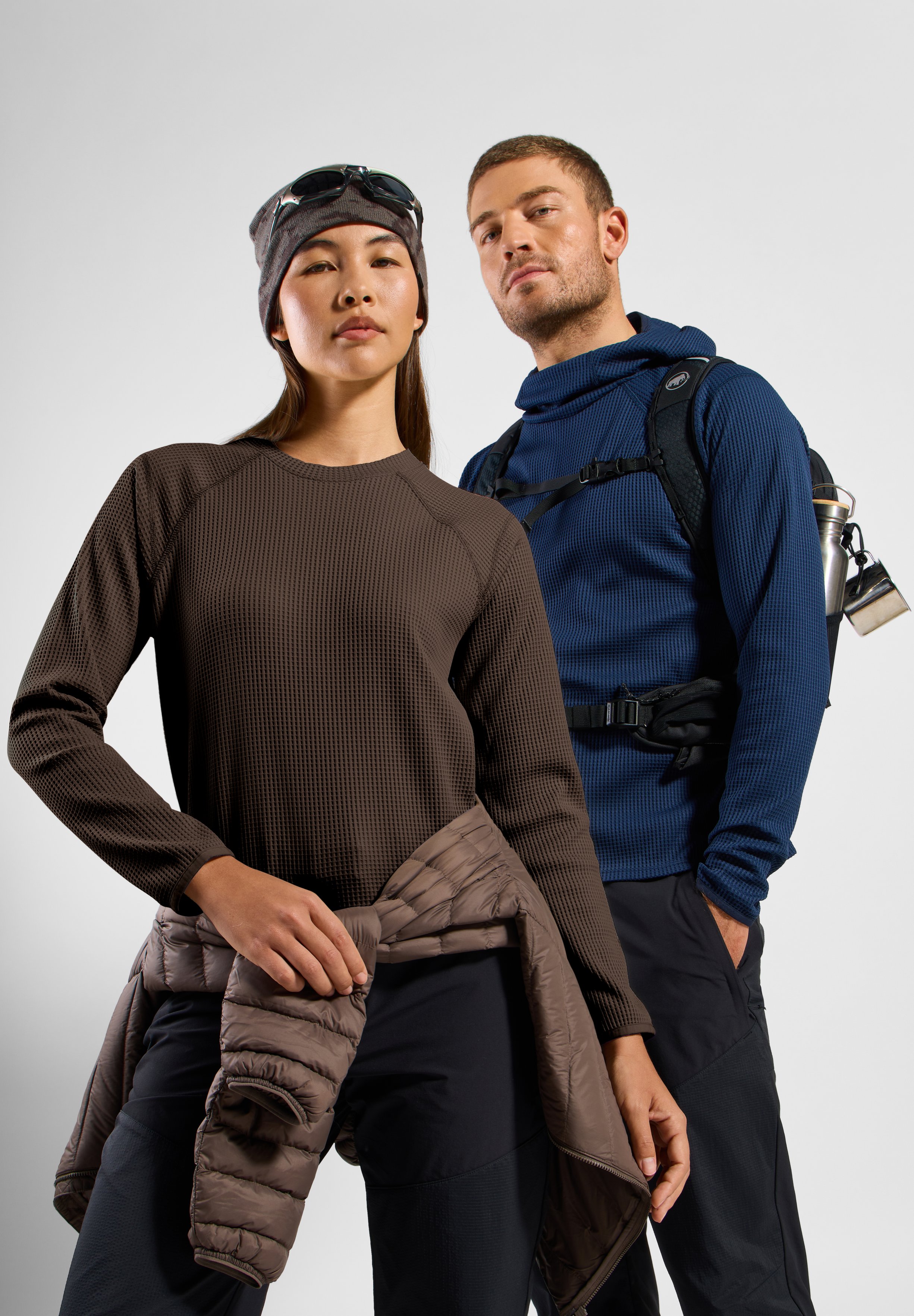 Woman in brown top, beanie and tied jacket, beside a man in a blue hoodie and backpack. Both model outdoor hiking apparel.