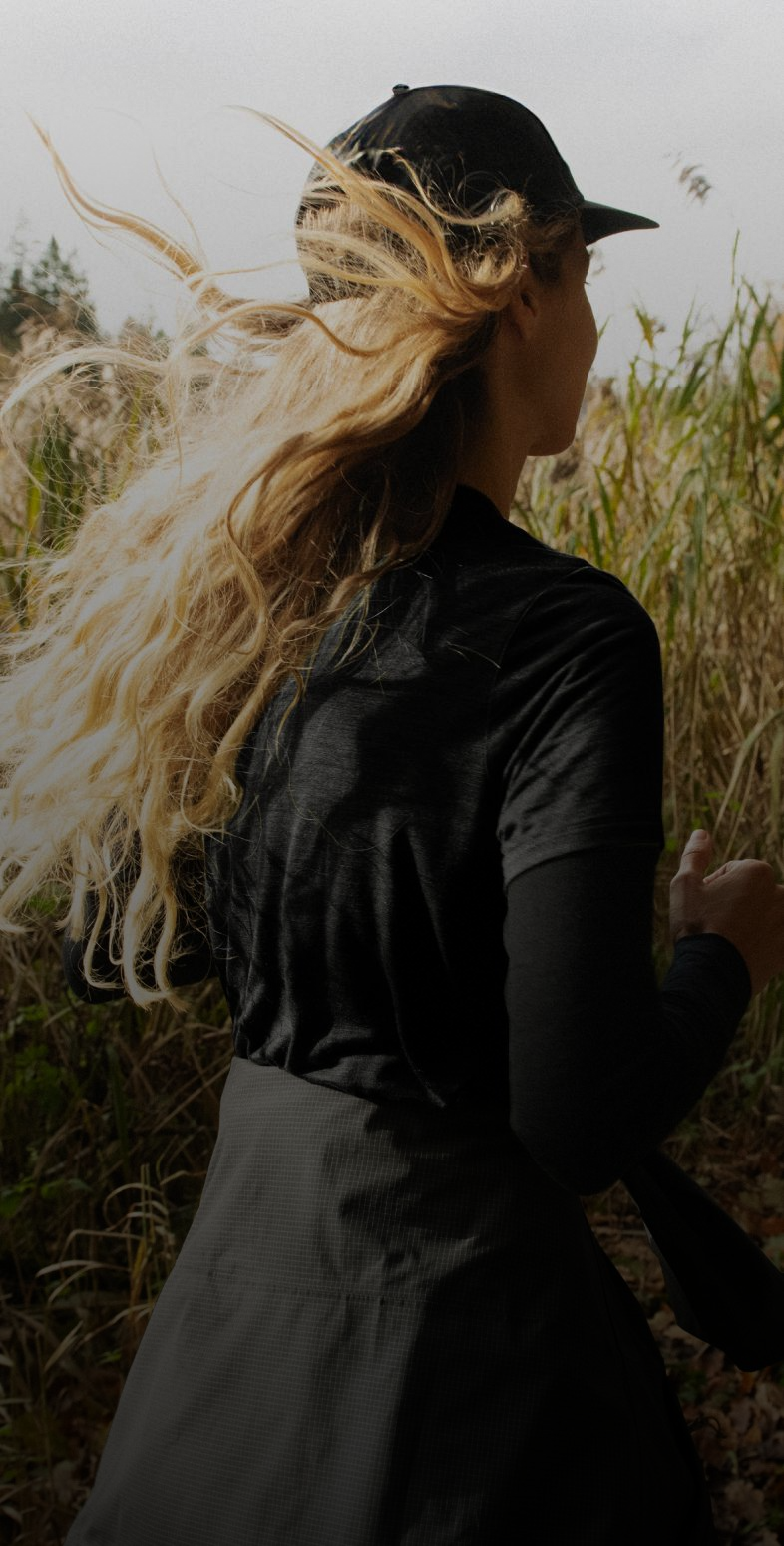 Woman with flowing blonde hair running through tall grass, wearing a black cap, black top, and grey grid skirt.