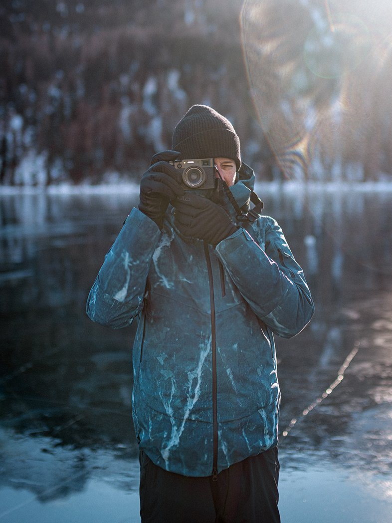 Filip Zuan in blauer Winterjacke und Beanie beim Fotografieren auf einem zugefrorenen See, die Sonne strahlt ins Bild, im Hintergrund ein verschneiter Wald.