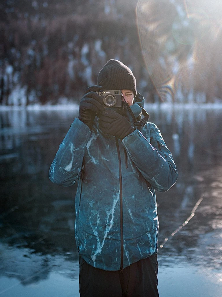 Filip Zuan in blauer Winterjacke und Beanie beim Fotografieren auf einem zugefrorenen See, die Sonne strahlt ins Bild, im Hintergrund ein verschneiter Wald.