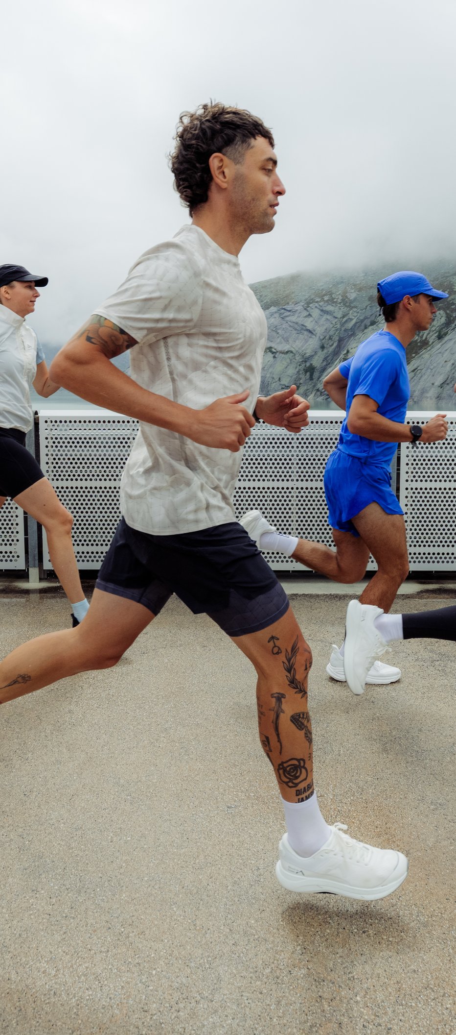Three runners in sportswear jogging on a paved path past a white fence, with foggy mountains in the background.