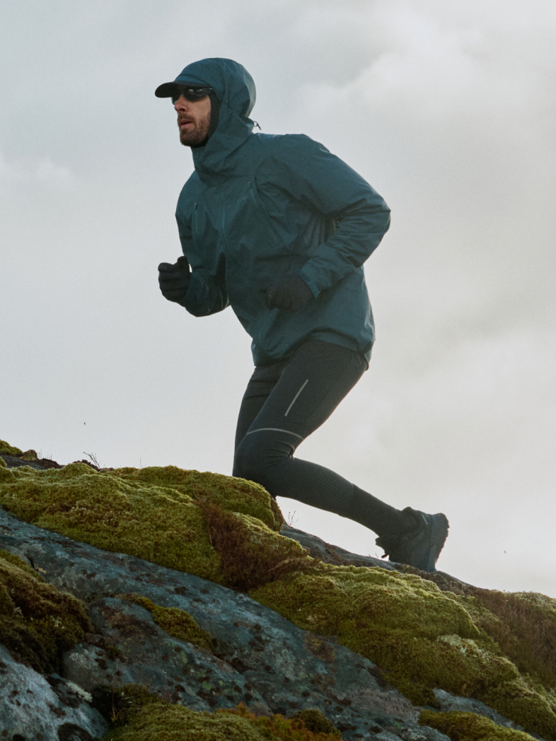 Mann läuft über bemoste Felsen in einer petrolblauen Kapuzenjacke, schwarzer Mütze, Sonnenbrille und dunklen Leggings vor einem bedeckten Himmel.