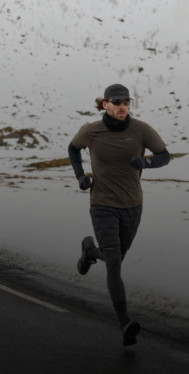 Ein Mann in braunem T-Shirt, schwarzer Leggings, Cap und Sonnenbrille läuft auf einer Straße durch eine verschneite Berglandschaft.