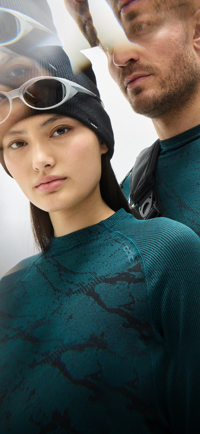 Woman in reflective orange ski goggles and black top stands by a man in a teal patterned shirt holding a black backpack.