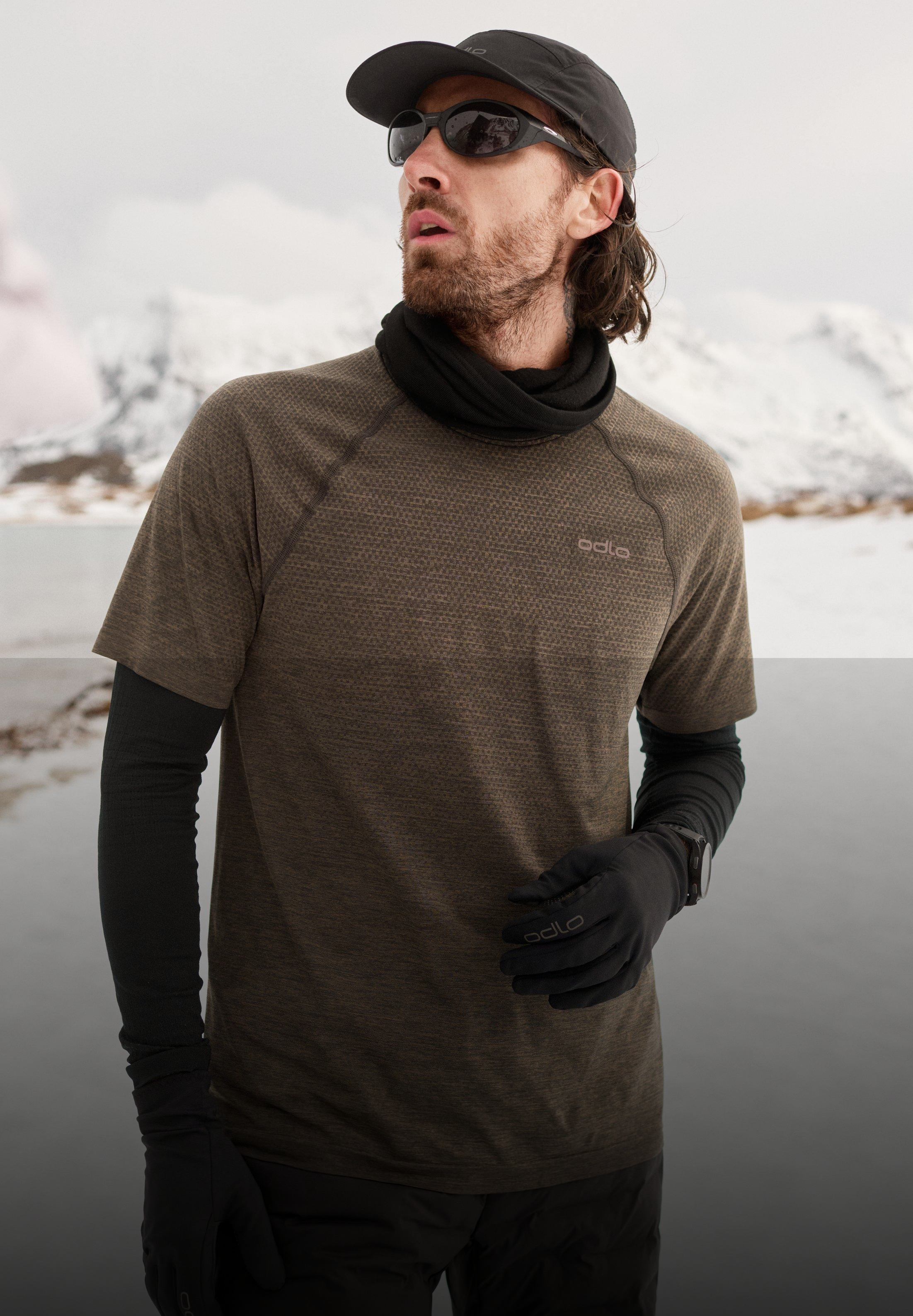 Mann, der auf einer nassen Straße mit verschneitem Hintergrund läuft und ein braunes T-Shirt, eine schwarze Shorts, eine Leggings, eine Cap und eine Sonnenbrille trägt.