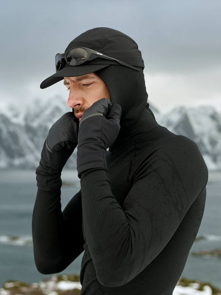 Mann in schwarzer Thermobekleidung und Handschuhen richtet seine Kapuze und Mütze mit Sonnenbrille, während er an eisigem Wasser steht, im Hintergrund schneebedeckte Berge.