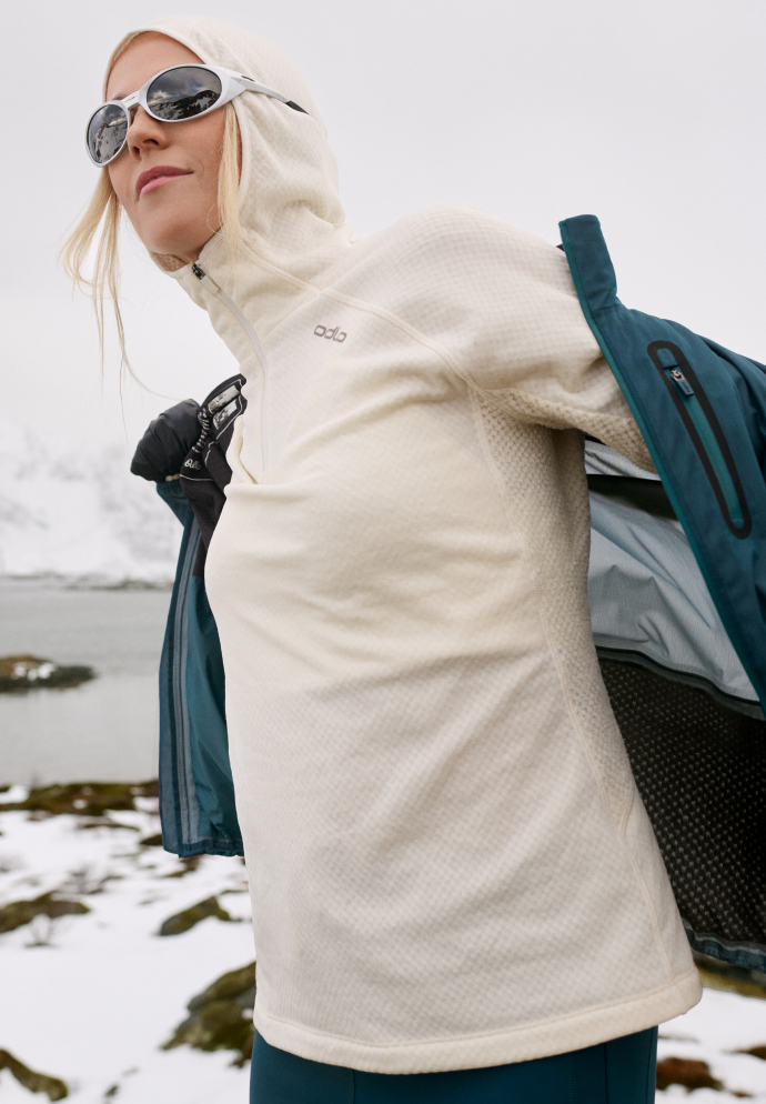 Blonde woman in a snowy landscape wearing sunglasses and a cream hooded mid-layer, taking off a teal jacket.
