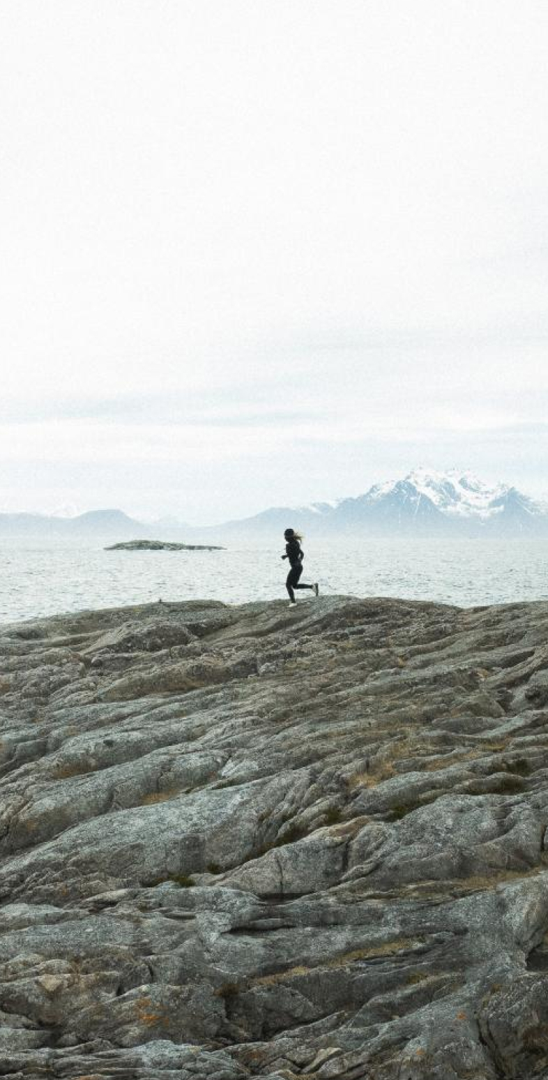 A person running on a rocky shore by the sea, with distant snow-capped mountains under a cloudy grey sky.