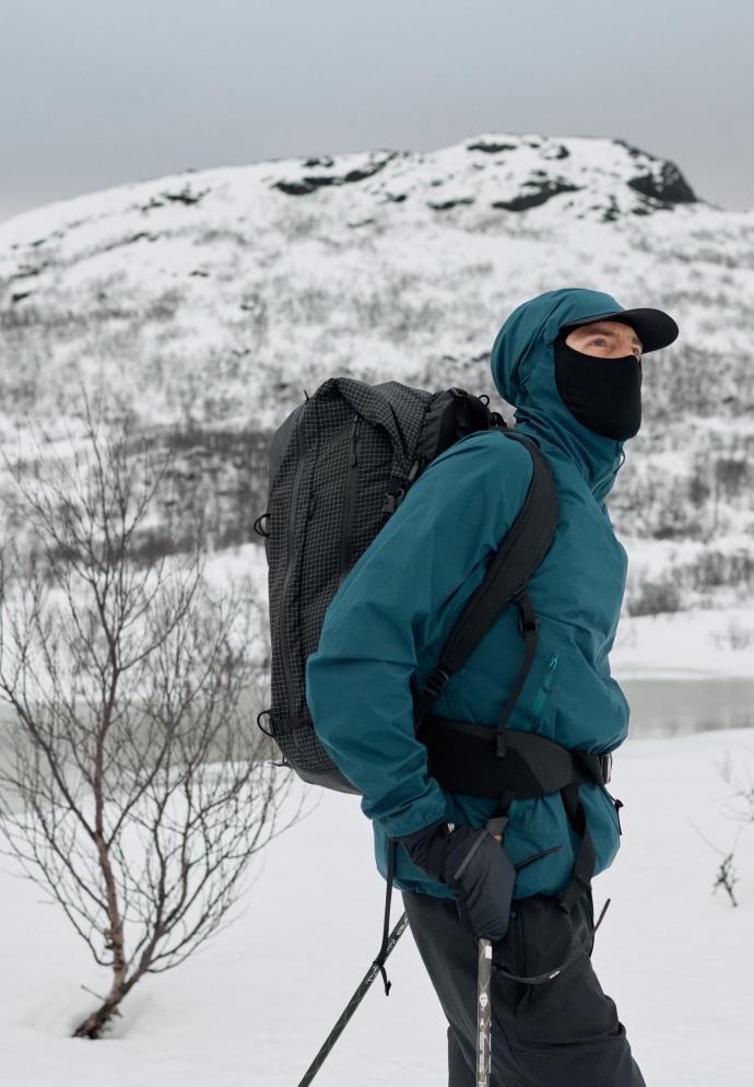 Person im Profil, trägt eine nasse, türkisfarbene Jacke mit Kapuze, eine schwarze Kappe, eine Sonnenbrille und eine Gesichtsmaske in verschneiter Umgebung.