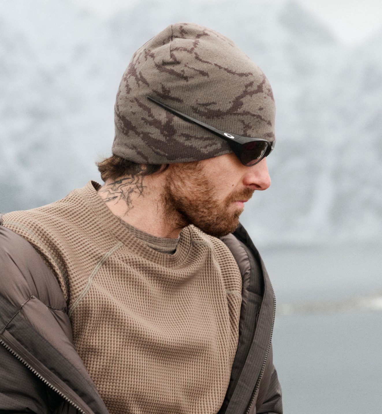 Profiel van een man met baard en nektattoo, bruine camobeanie, zonnebril en beige shirt tegen een besneeuwd berglandschap.