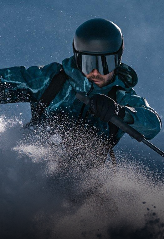 Een skiër in een groenblauwe jas en met een zwarte helm carvet door diepe poedersneeuw en laat sneeuw opstuiven tegen een donkerblauwe lucht.