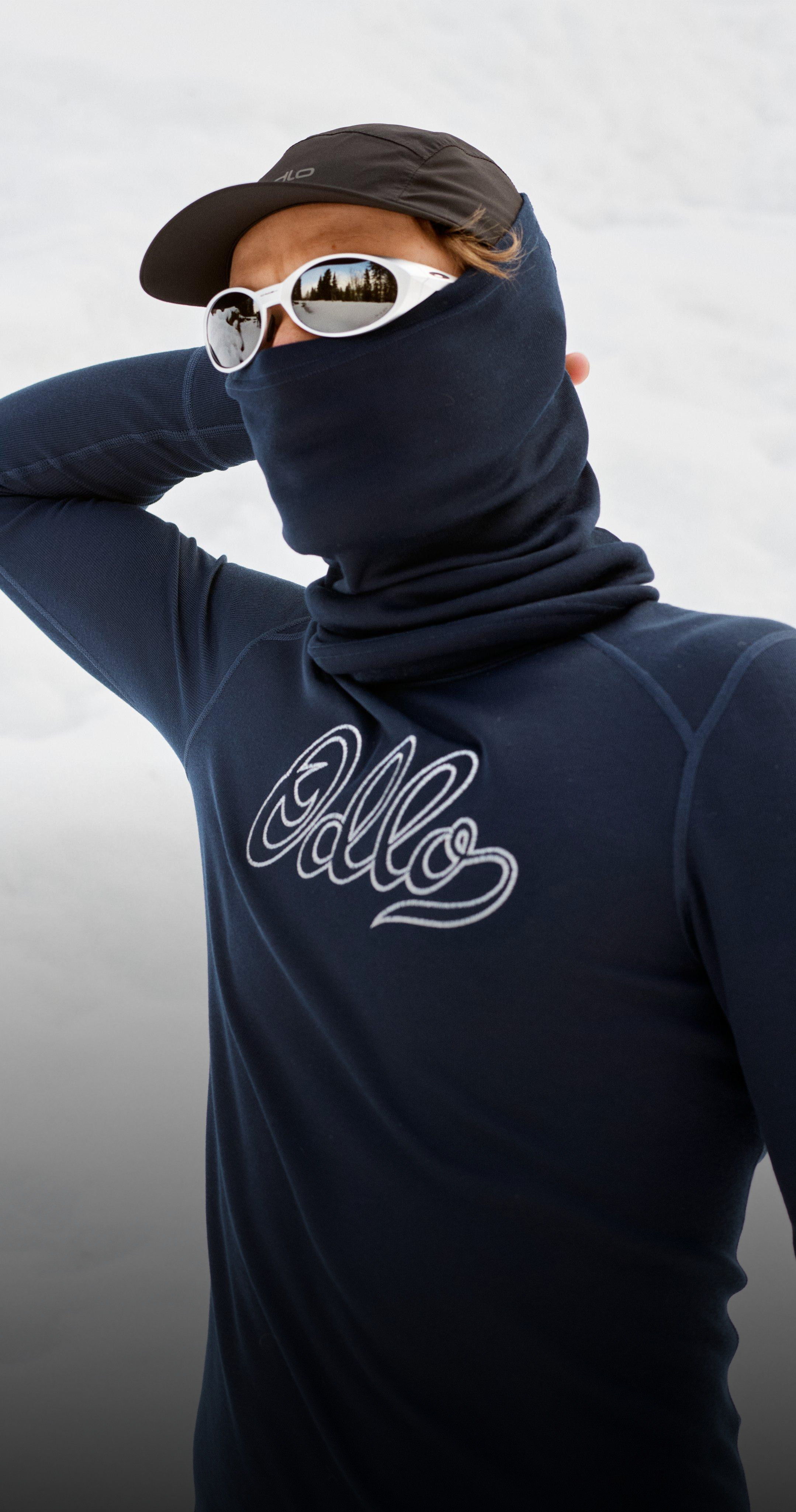 Man in marineblauwe base layer met "Odlo"-logo, gezicht bedekt door een nekwarmer, met een witte zonnebril en een pet, staand in de sneeuw.