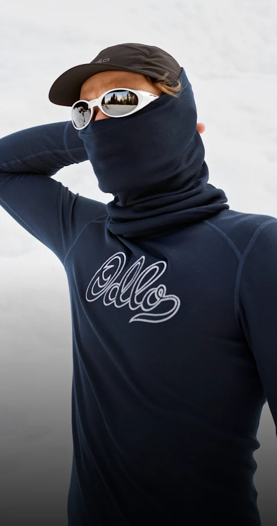 Man in marineblauwe base layer met "Odlo"-logo, gezicht bedekt door een nekwarmer, met een witte zonnebril en een pet, staand in de sneeuw.