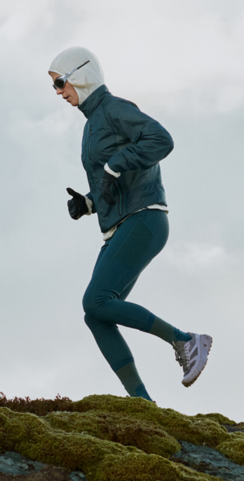 Un runner in giacca color verde acqua, leggings, cappuccio bianco e occhiali da sole corre su un terreno verde muschio sotto un cielo nuvoloso.