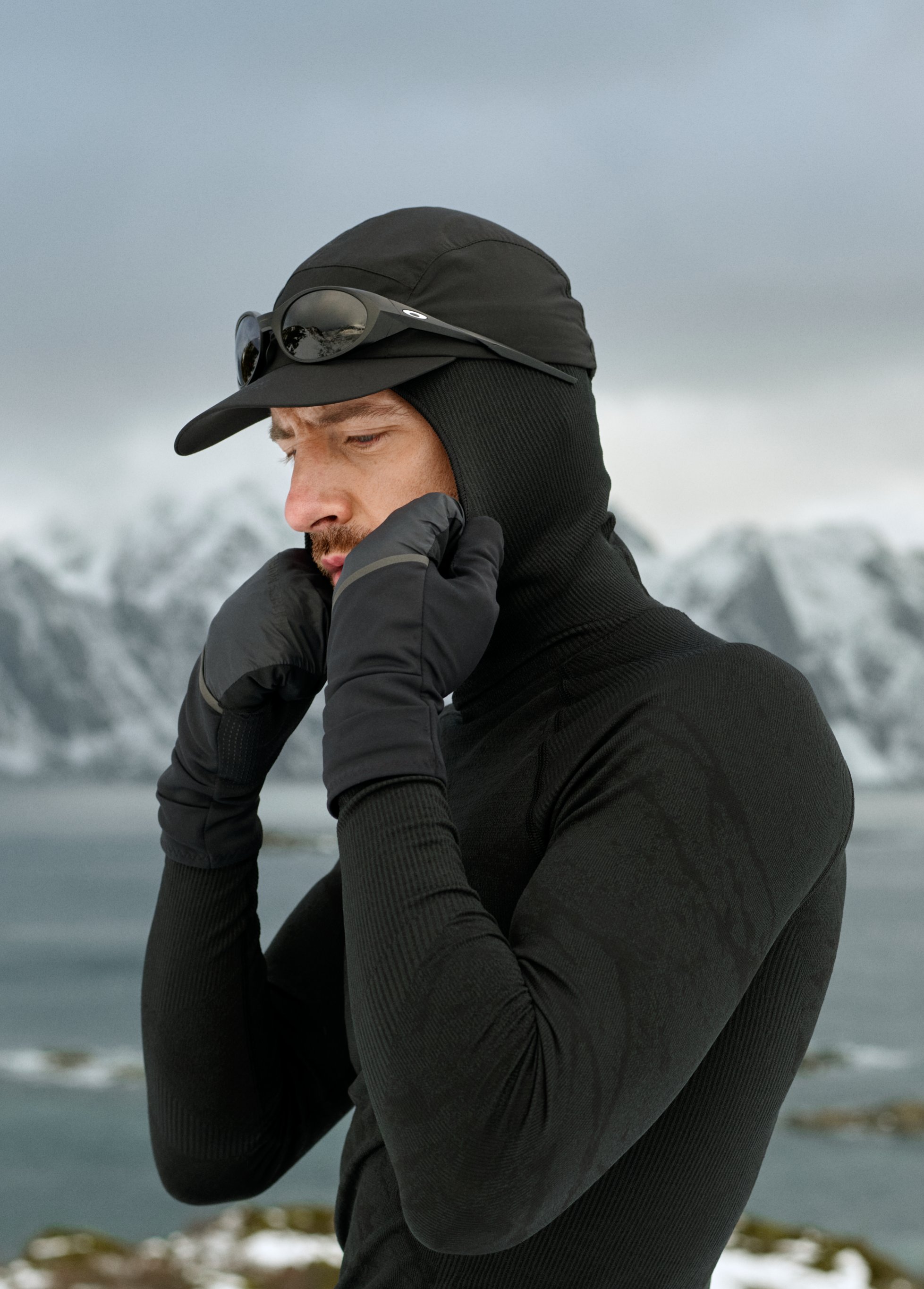 Uomo in abbigliamento termico nero, berretto e guanti si sistema il cappuccio con montagne innevate e acqua sullo sfondo.