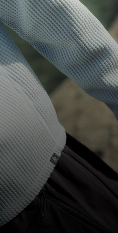 Close-up van een lichtblauwe longsleeve met wafelstructuur en een logolabel bij de zoom, gedragen met een zwarte broek.