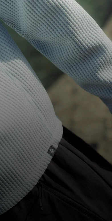 Close-up van een lichtblauwe longsleeve met wafelstructuur en een logolabel bij de zoom, gedragen met een zwarte broek.