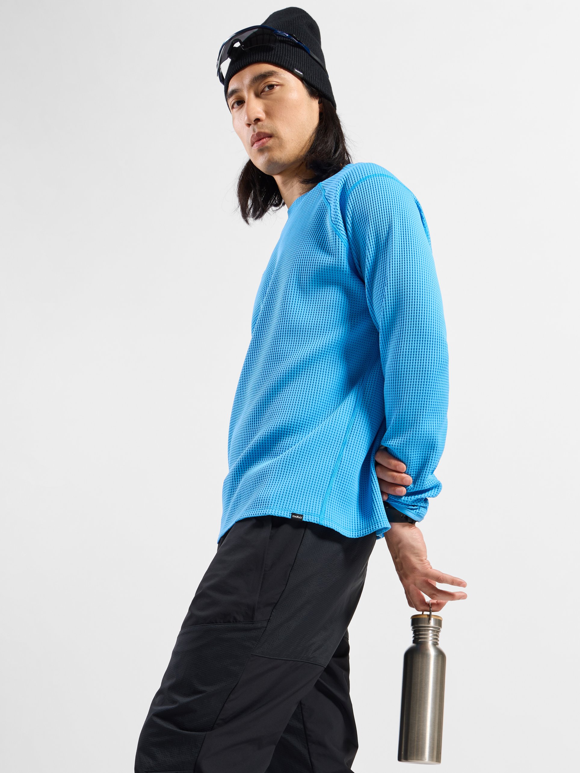 Long-haired man in a blue shirt, black pants and beanie, looking back while holding a silver water bottle behind him.