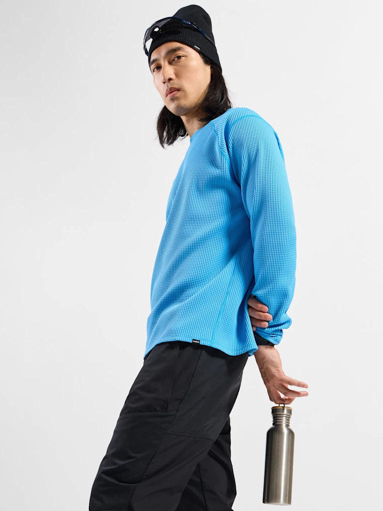 Long-haired man in a blue shirt, black pants and beanie, looking back while holding a silver water bottle behind him.