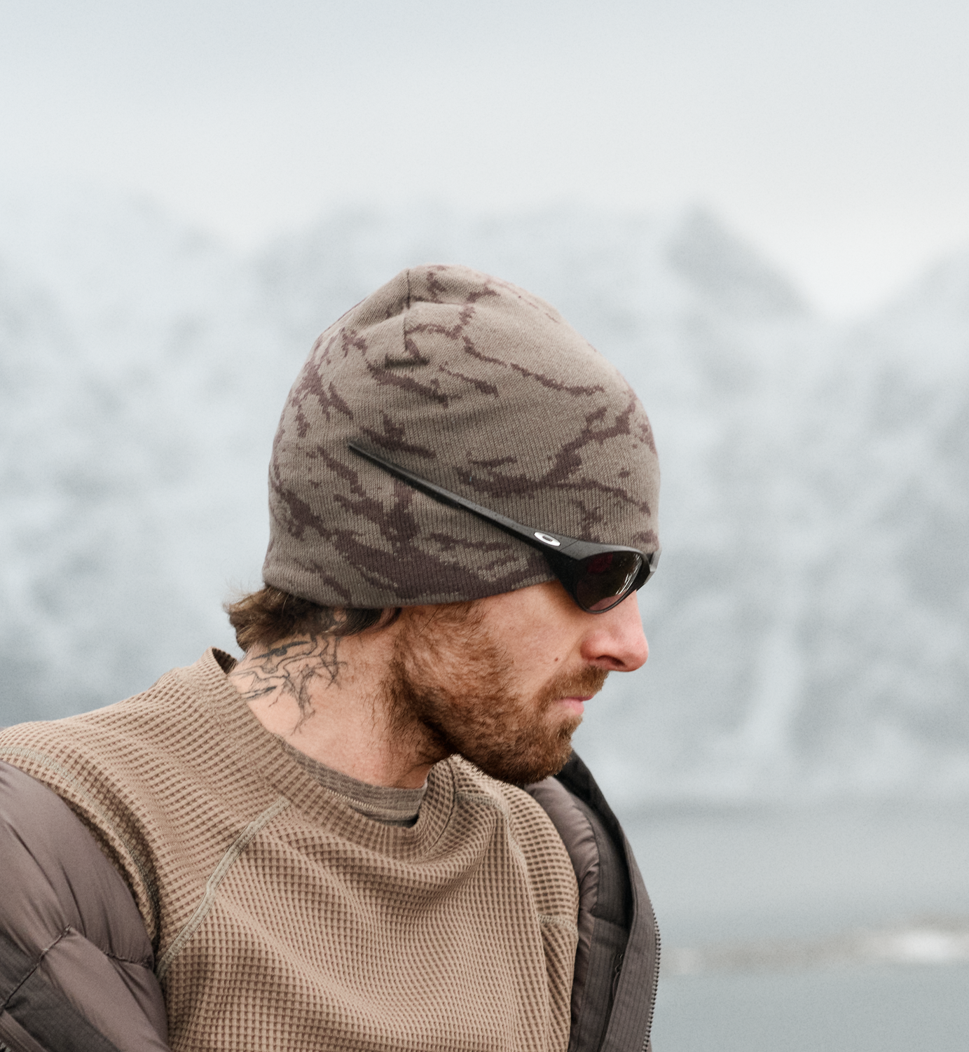 Profiel van een man met baard en nektattoo, camouflagemuts en zonnebril tegen een wazige achtergrond van besneeuwde bergen.