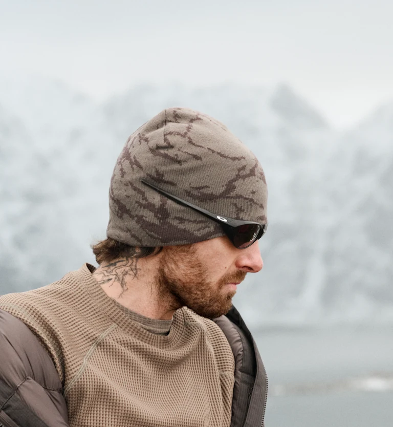 Profiel van een man met baard en nektattoo, camouflagemuts en zonnebril tegen een wazige achtergrond van besneeuwde bergen.