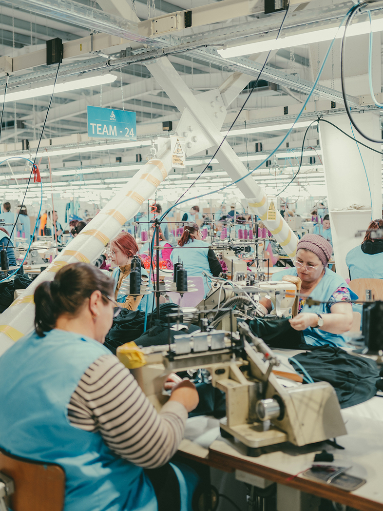 Viele Frauen in hellblauen Westen arbeiten in einer hellen, großen Bekleidungsfabrik an Nähmaschinen und verarbeiten dunklen Stoff.