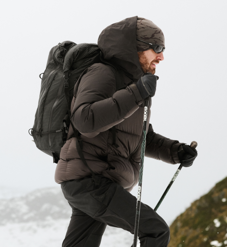 Ein Mann mit Bart, in dunkelbrauner Daunenjacke, mit schwarzem Rucksack und Sonnenbrille, wandert mit Trekkingstöcken auf einem verschneiten Berg.