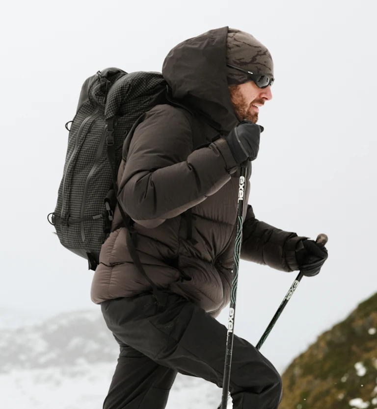 Ein Mann mit Bart, in dunkelbrauner Daunenjacke, mit schwarzem Rucksack und Sonnenbrille, wandert mit Trekkingstöcken auf einem verschneiten Berg.