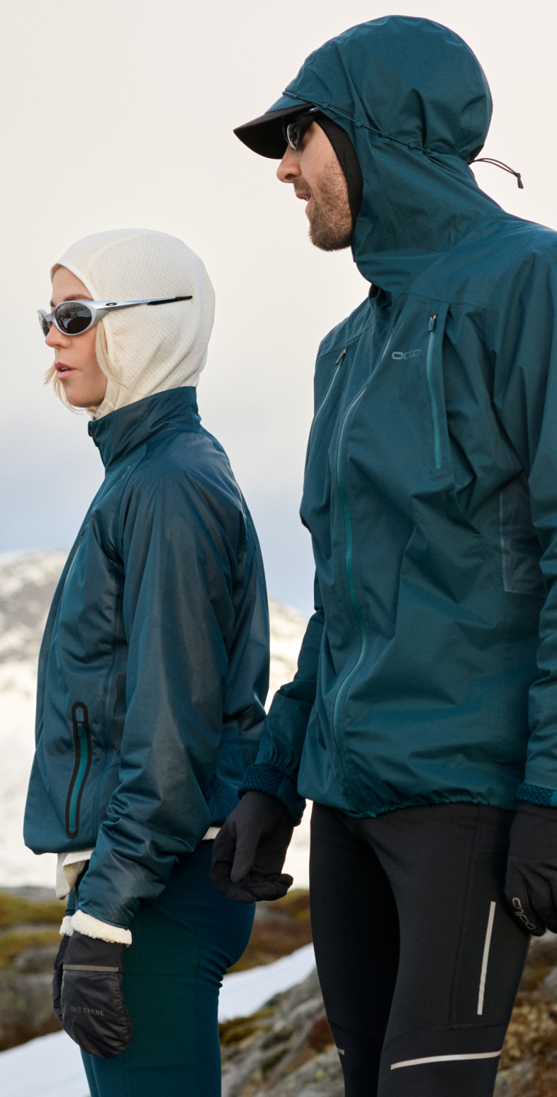 A man and woman in teal hooded jackets and sunglasses look left, standing in a snowy mountain landscape.