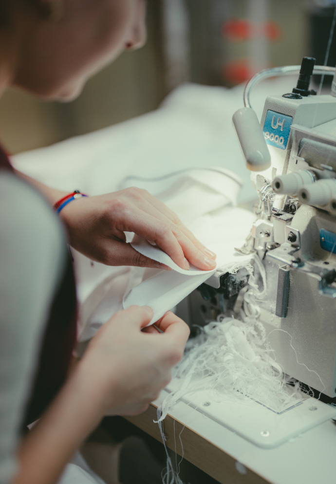 A person's hands guiding white fabric through an industrial sewing machine, surrounded by white thread scraps.