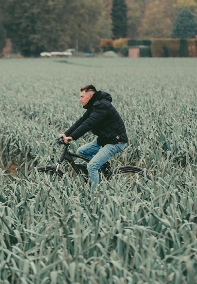 Ein Mann in schwarzer Jacke und blauen Jeans fährt mit einem schwarzen Fahrrad durch ein Feld mit hohem, grünem Getreide.