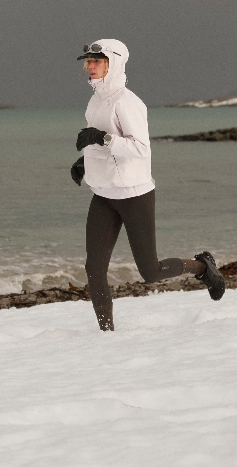 Une femme en veste blanche à capuche et legging noir court sur une plage enneigée au bord de l'océan, sous un ciel sombre et orageux.