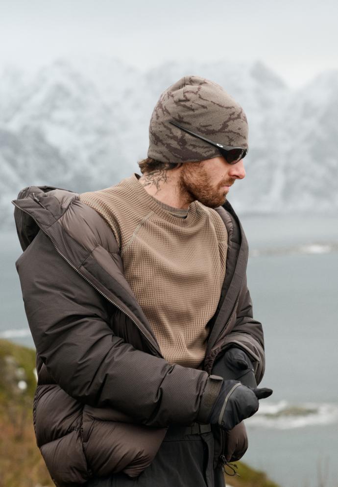 Mann mit Beanie im Camo-Muster, Mid Layer mit Gitterstruktur und brauner Steppjacke richtet seine Handschuhe vor einem verschneiten Berg- und Wasser-Hintergrund.