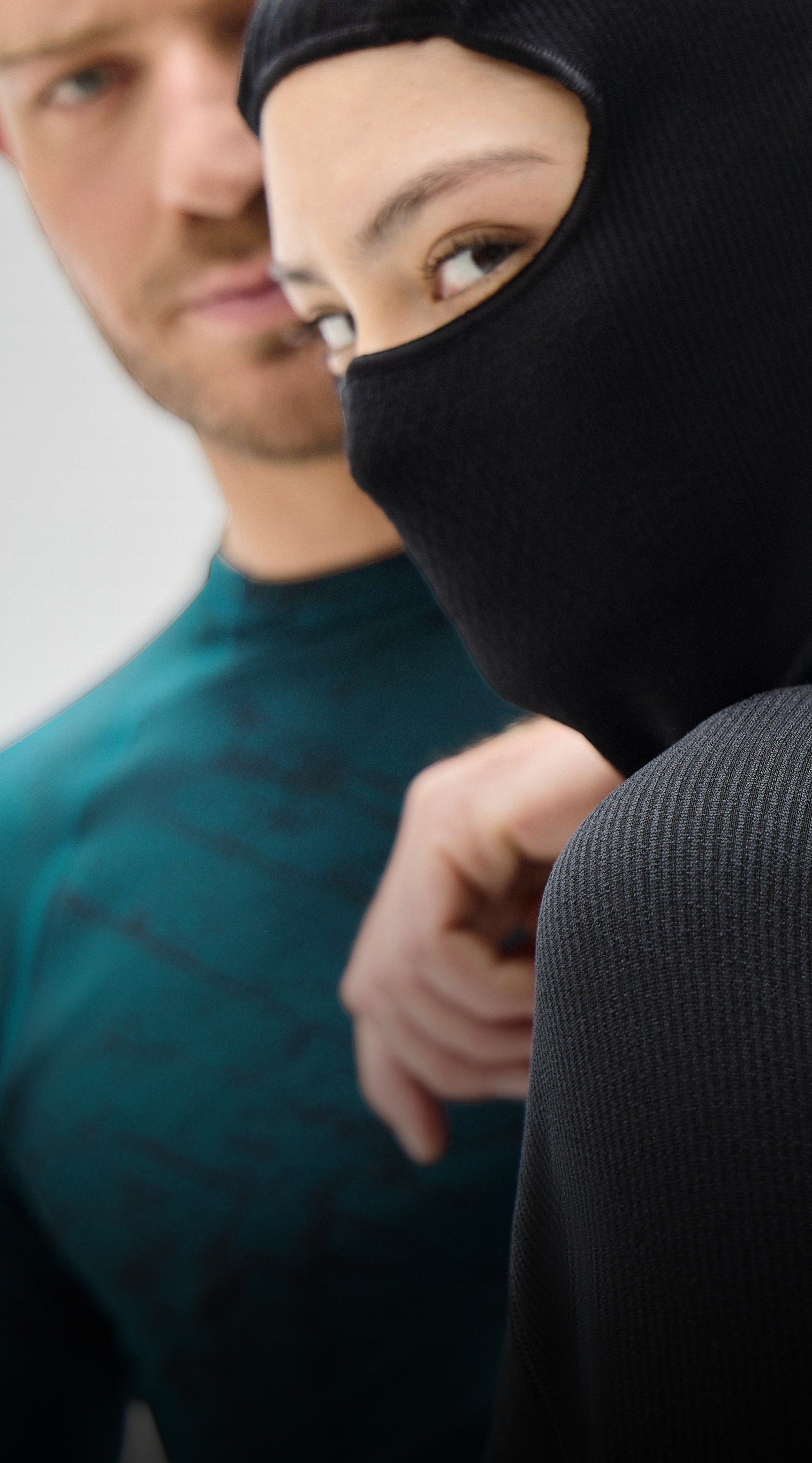 Nahaufnahme von zwei Personen in Thermo-Bekleidung: Eine trägt eine schwarze Sturmhaube, die andere ein türkisfarbenes Langarmshirt. Beide blicken in die Kamera.