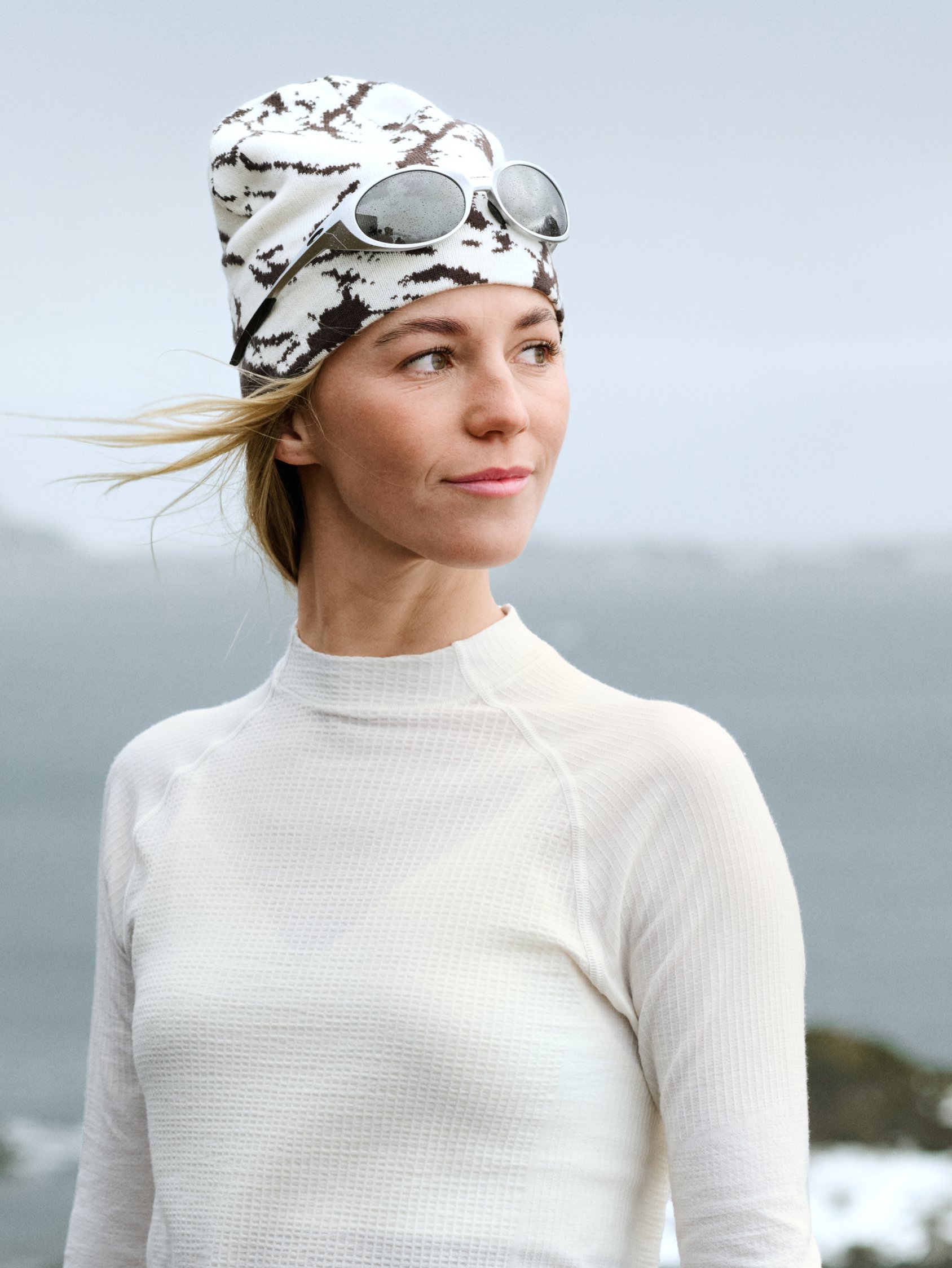 Frau in weißem Thermoshirt und gemusterter Beanie mit Skibrille, die an einem windigen, bewölkten Tag draußen an der Küste nach rechts schaut.