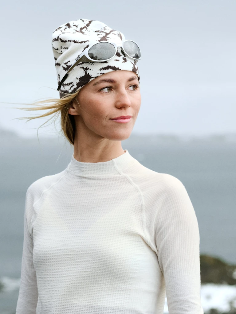 Frau in weißem Thermoshirt und gemusterter Beanie mit Skibrille, die an einem windigen, bewölkten Tag draußen an der Küste nach rechts schaut.
