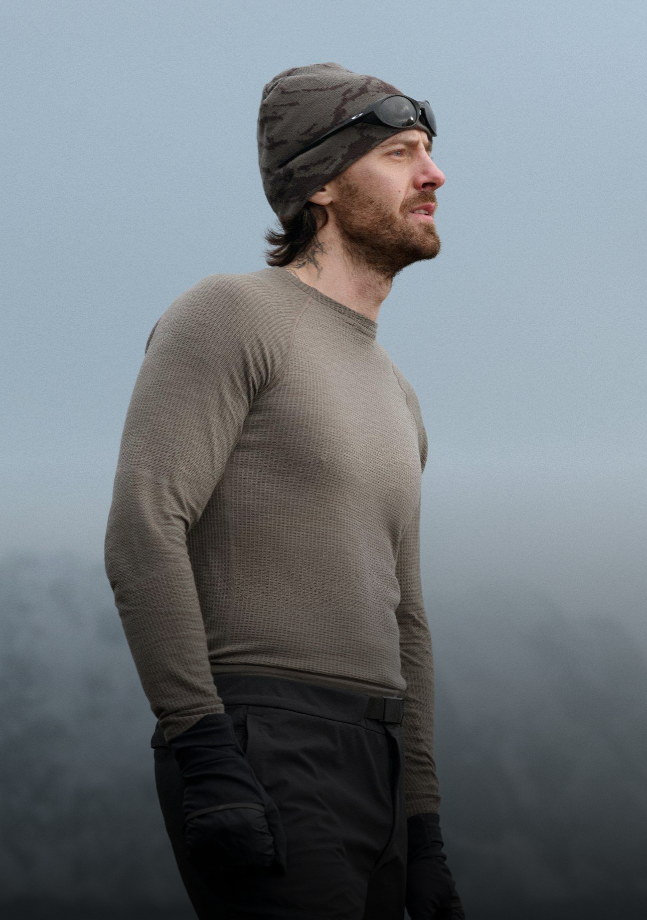 Bärtiger Mann in einem braunen Performance Wool Thermoshirt, Camouflage-Beanie und schwarzen Handschuhen blickt zur Seite vor einem nebligen Bergpanorama.