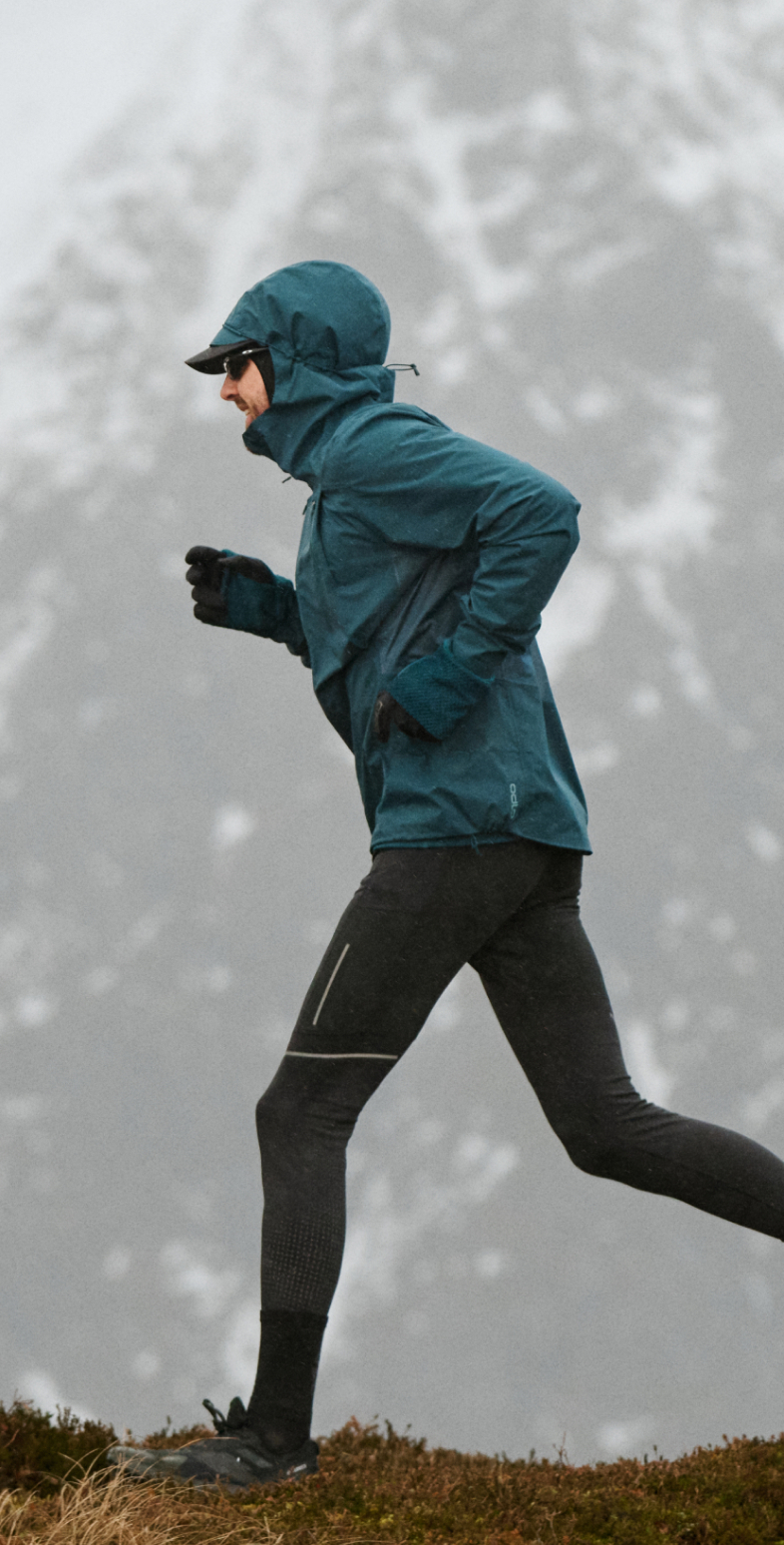 Een hardloper in een blauw jack en zwarte legging jogt over een grazige bergkam met besneeuwde, mistige bergen op de achtergrond.