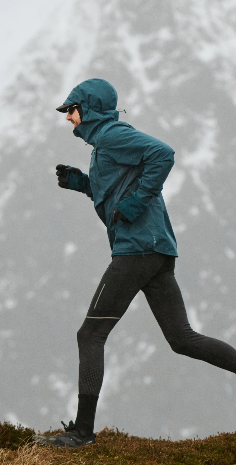 A runner in a blue jacket and black leggings jogs on a grassy ridge with snowy, foggy mountains in the background.
