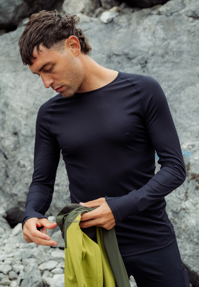 Man in a dark blue long-sleeve top stands against grey rocks, looking down while holding a green and yellow garment.