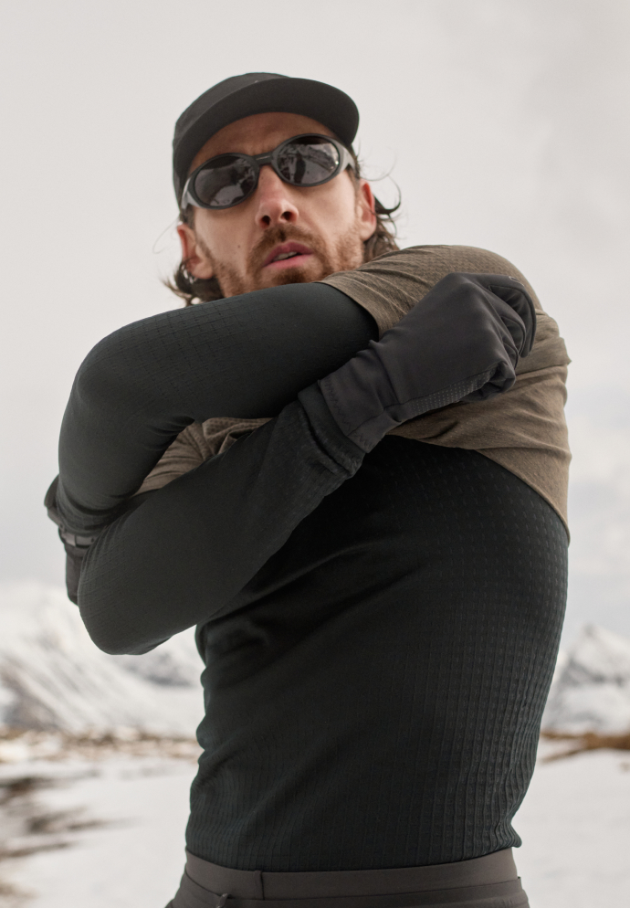 Bärtiger Mann mit Mütze und Sonnenbrille, der sich ein olivfarbenes T-Shirt über ein schwarzes Langarmshirt zieht – in einer verschneiten Berglandschaft.