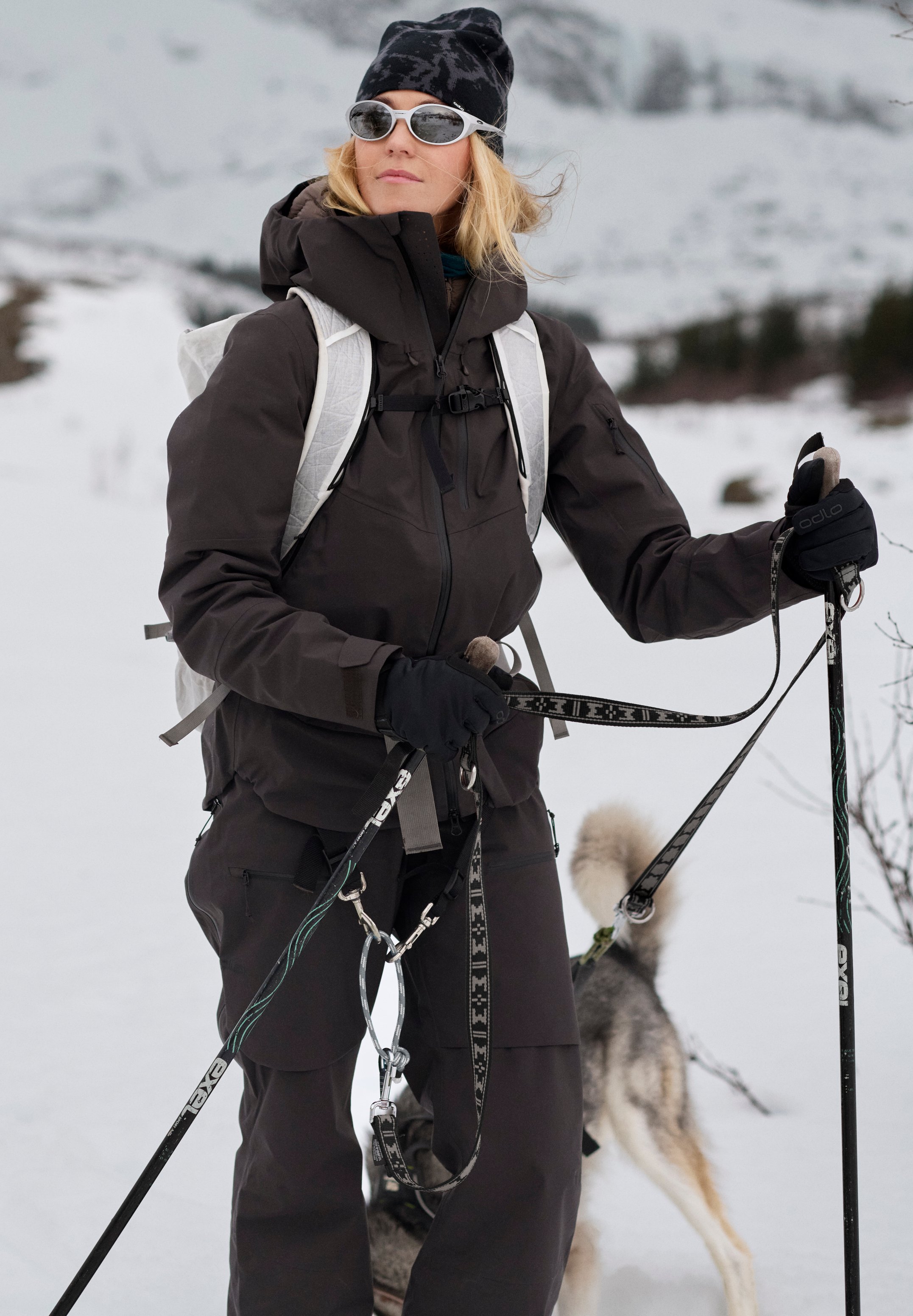 Vrouw in donkere winterkleding, met muts, zonnebril en witte rugzak, staat in de sneeuw met skistokken in haar handen. Achter haar staat een husky.