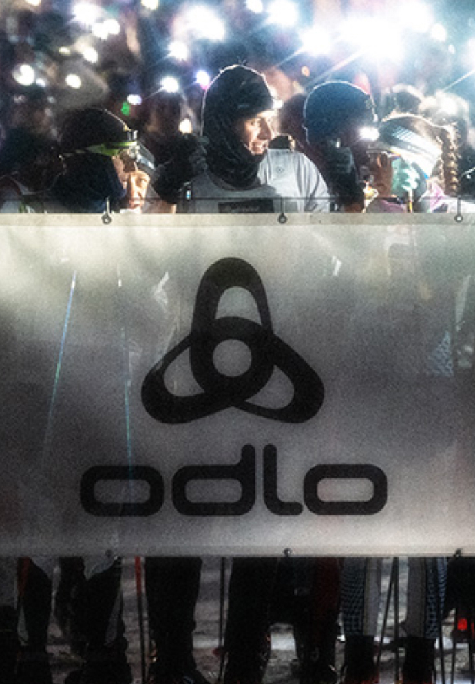 Cross-country skiers with bright headlamps wait behind a white Odlo banner at a night race start line.