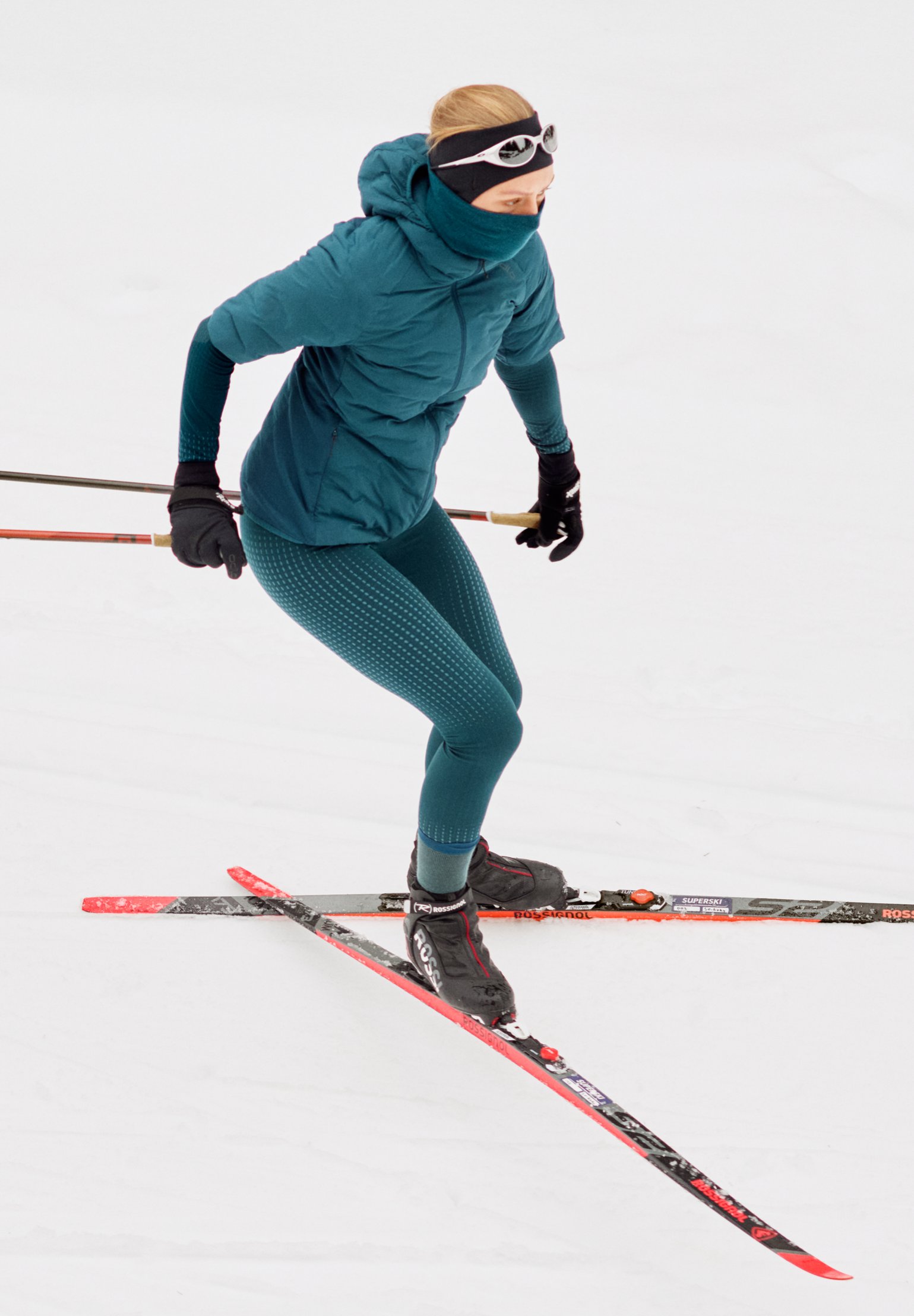 Une femme fait du ski de fond dans la neige, vêtue d'une veste bleu-vert, d'un legging à motifs et d'un tour de cou, chaussée de skis rouges.