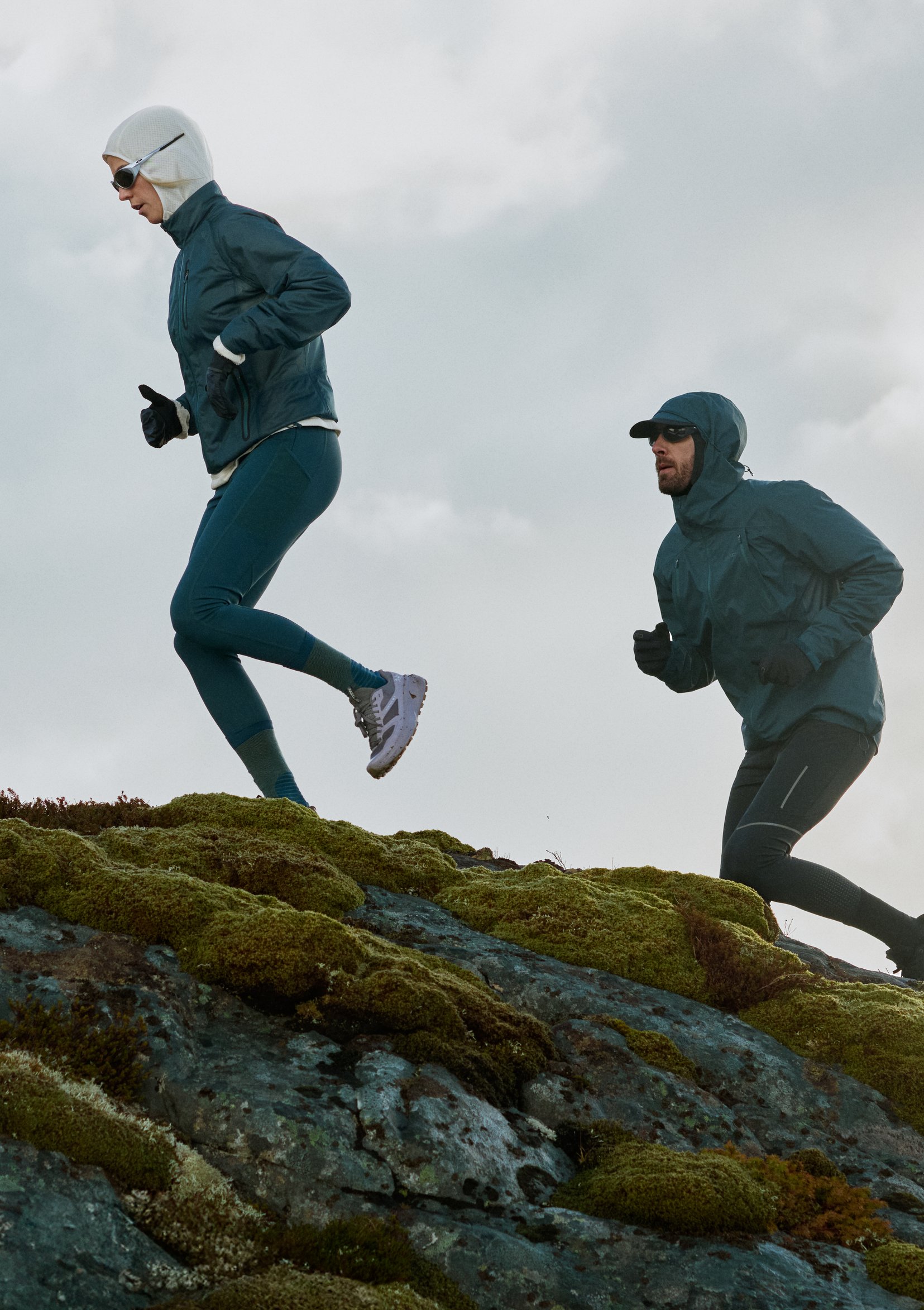 Zwei Personen in dunkelblauer Sportbekleidung laufen auf moosbedeckten Felsen unter einem bewölkten Himmel bergauf.